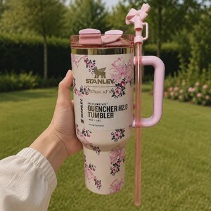 May include: A cream-colored Stanley Quencher H2.0 tumbler with a floral and bow pattern, featuring a pink handle, a matching pink straw with a bow topper, and a clear lid. The label reads Stanley, LoveShackFancy, and The Flowstate Quencher H2.0 40 oz.