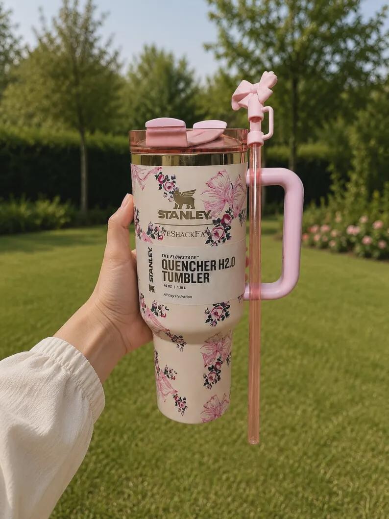 May include: A cream-colored Stanley Quencher H2.0 tumbler with a floral and bow pattern, featuring a pink handle, a matching pink straw with a bow topper, and a clear lid. The label reads Stanley, LoveShackFancy, and The Flowstate Quencher H2.0 40 oz.