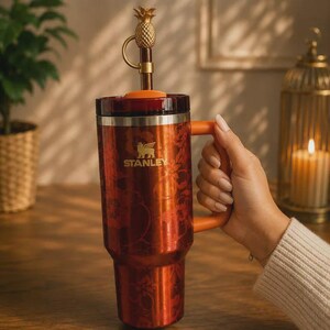 May include: A tall, metallic orange Stanley tumbler featuring a floral pattern, a sturdy orange handle, and a lid topped with a decorative gold-colored pineapple finial. The brand name Stanley is printed in gold on the front of the insulated cup.