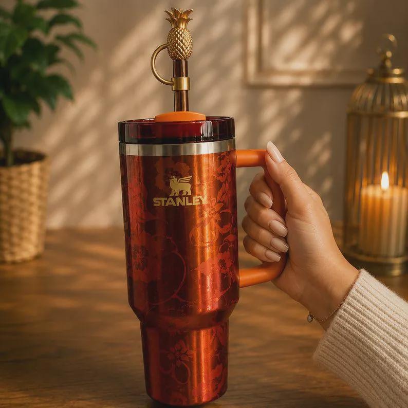 May include: A tall, metallic orange Stanley tumbler featuring a floral pattern, a sturdy orange handle, and a lid topped with a decorative gold-colored pineapple finial. The brand name Stanley is printed in gold on the front of the insulated cup.