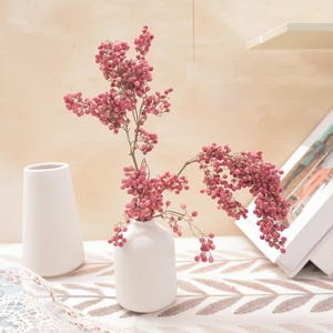 Peut inclure: Deux vases en céramique blanche, l'un grand et conique, l'autre court et arrondi, contiennent des branches de baies roses. Les baies sont regroupées le long des tiges. Les vases sont posés sur une surface à motifs, avec un fond neutre.