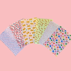 May include: A set of five rectangular sheets of patterned paper arranged in a fan shape. The patterns include pink strawberries, orange fish shapes, light green dinosaur silhouettes, white line art, and colorful rainbows with hearts and clovers.