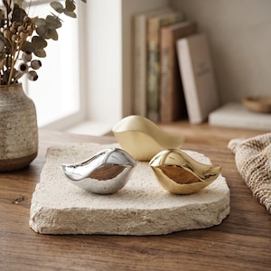 May include: Three bird-shaped figurines arranged on a textured, light-colored stone slab. One figurine is polished silver, one is polished gold, and one is a matte gold finish. The birds have smooth, rounded bodies and minimalist, abstract shapes.