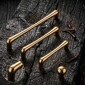 May include: Three polished gold-toned metal cabinet pulls of varying lengths and one matching round cabinet knob. The hardware features a smooth, curved design with rounded ends. Several small metal screws are scattered nearby.