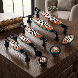 May include: A collection of cabinet hardware including four arched drawer pulls and three knobs, each featuring a black metal base and a central ceramic insert with a starburst pattern in orange, white, and black against a teal marbled background.