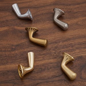 May include: Five metal wall hooks in various finishes, including polished chrome, brushed nickel, matte gold, polished brass, and satin gold. Each hook features a flared, fan-shaped base that tapers into a curved, cylindrical arm with a rounded end.
