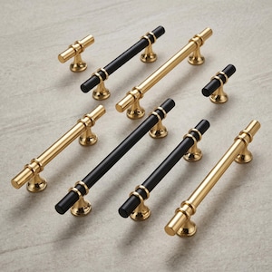 Könnte beinhalten: Eine Sammlung von Schrankbeschlägen bestehend aus goldfarbenen Metallgriffen und Knöpfen mit mattschwarzen zylindrischen Mittelteilen. Das Set enthält Stangengriffe in verschiedenen Längen sowie kleine T-förmige Knöpfe mit polierten Goldbasen.