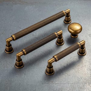 May include: A collection of antique brass hardware including three cylindrical cabinet pulls with a textured knurled surface and one round cabinet knob. Each piece features a weathered metallic finish with dark highlights and sturdy mounting posts.