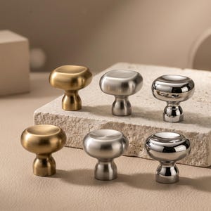 May include: Six cabinet knobs arranged in two rows, featuring three distinct finishes: brushed gold, brushed silver, and polished chrome. Each knob has a rounded, mushroom-like top and a tapered base, showcasing a smooth, metallic surface texture.