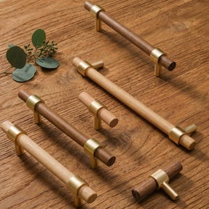 May include: A collection of cabinet hardware featuring cylindrical wooden bars in light and dark wood tones, accented with brushed gold metal mounting posts. The set includes various lengths of drawer pulls and T-shaped knobs for furniture.