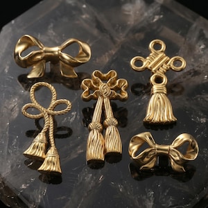 May include: A collection of five gold-toned metal charms featuring two smooth bows, two rope-textured tassels with looped tops, and one ornate knot design with a single tassel. Each piece has a polished, reflective finish and intricate detailing.