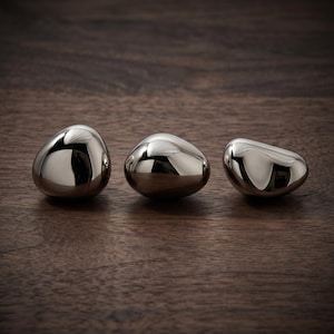 Puede incluir: Tres objetos metálicos lisos y pulidos con forma de piedras de río redondeadas. Cada pieza presenta una superficie plateada altamente reflectante, similar a un espejo, que captura la luz y el entorno, creando reflejos curvos.