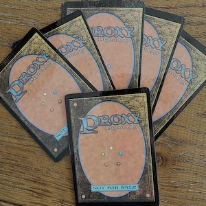 Puede incluir: Una colección de cartas de juego rectangulares con un centro ovalado de color tostado y cinco pequeños puntos de colores dispuestos en forma de pentágono. Las cartas muestran el texto Proxy Playtest Copy y Not For Sale en azul.