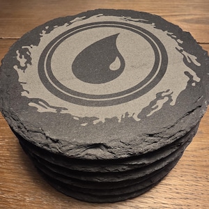May include: A stack of circular black slate coasters with rough, uneven edges. The top coaster features a laser-engraved design of a central water droplet icon enclosed within two concentric circles, surrounded by a stylized, irregular splash pattern.