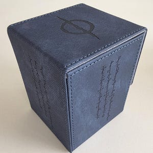 May include: A dark blue, textured, cube-shaped storage box with stitched edges. The top surface features a circular symbol with a horizontal line, while the front panel displays three vertical columns of etched, runic-style characters in black.