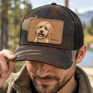 Op de afbeelding: Een camouflagekleurige truckerpet met een rechthoekige, lichtbruine leren patch aan de voorzijde. De patch toont een gedetailleerde illustratie van een goudkleurige hond met krullend haar en de naam Willow in zwarte letters rechtsonder.