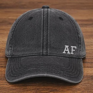 May include: A charcoal grey baseball cap with a washed, distressed finish. The front panel features the letters AF embroidered in white thread. The cap has a curved brim, a structured crown with stitched seams, and a button at the top centre.