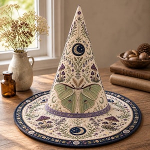 May include: A conical hat featuring cream-colored fabric with intricate embroidery of a large pale green moth, purple mushrooms, and celestial moon motifs. The design includes a wide brim with a dark blue border and a purple band at the base.