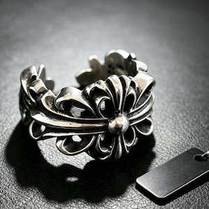 May include: A silver-toned metal ring featuring an open-ended band with a prominent, symmetrical floral-inspired motif. The design includes intricate, curved cutouts and a polished central sphere, showcasing a darkened, oxidised finish in the recessed areas.