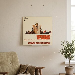Könnte beinhalten: Ein quadratisches Schallplattencover mit cremefarbenem Hintergrund und einer zentralen Illustration einer Person mit Hut und Stiefeln, flankiert von zwei großen Brotscheiben. Der Text lautet Mein Name ist Nobody, Original Film-Soundtrack und Ennio Morricone.
