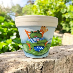 May include: A ceramic flower pot with a light blue background and a cream-colored rim. The pot features a cartoon frog wearing a crown, striped shirt, and blue swim trunks, relaxing in a chair and holding a drink. The frog is painted in bright green.