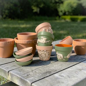 May include: A collection of terracotta and green ceramic flower pots and saucers. Some pots feature embossed designs of a rabbit, sunflower, and turtle. The pots are arranged on a weathered wooden surface, with a blurred green background.