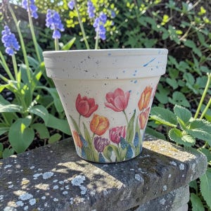 May include: A white ceramic flower pot with a painted floral design featuring red, orange, and purple tulips. The pot has blue paint splatters and sits on a stone wall, surrounded by greenery and blue flowers.