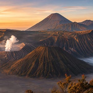 May include: A panoramic view of Mount Bromo and surrounding volcanic craters at sunrise. The landscape features a prominent smoking crater, rugged dark brown slopes, and a distant mountain peak under a soft orange and yellow sky with low clouds.