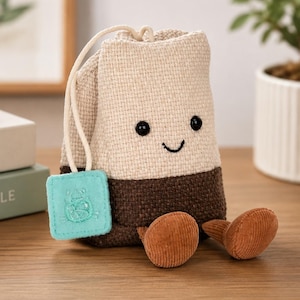 Puede incluir: Una caprichosa bolsita de té de peluche con una cara sonriente, ojos negros de botón y una base marrón. Tiene una etiqueta azul claro y dos piezas en forma de hongo de pana marrón. La bolsita de té está hecha de tela beige y marrón.