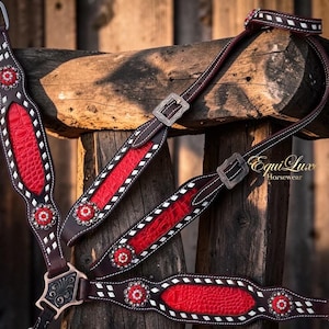 May include: A dark brown leather horse breast collar featuring bright red textured inlays, white stitched diamond patterns, and circular floral conchos with red and white stones. The hardware is metallic with engraved details and the text Equi Luxe.
