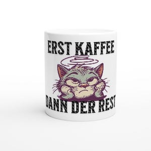 Könnte beinhalten: Weiße Keramik-Kaffeetasse mit einer Illustration einer grummeligen Katze und dem Text "ERST KAFFEE DANN DER REST". Die Katze ist grau und lila, mit gerunzelter Stirn und Pfoten unter dem Kinn.