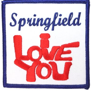 May include: A white embroidered patch with blue stitching around the edges. The patch features the text "Springfield" in blue cursive lettering and the text "Love You" in red stylized lettering.