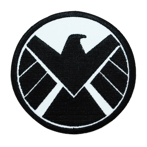 May include: Black and white embroidered patch featuring the S.H.I.E.L.D. logo. The logo is a circle with a black eagle inside and white lines radiating out from the center.