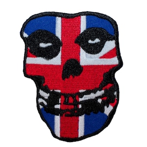 Può includere: Una toppa di teschio rosso, bianco e blu con il design della bandiera Union Jack. Il teschio è nero con occhi bianchi e una bocca nera.