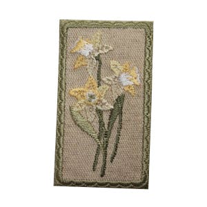 May include: A beige embroidered patch with a green border featuring three yellow daffodils with white centers.
