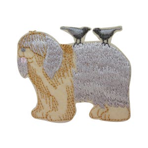May include: Embroidered patch of a fluffy, light brown dog with two black birds perched on its back. The dog has a long, shaggy coat and a sweet expression. The birds are facing opposite directions.