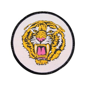 May include: A white circular patch with a black border features a yellow and black tiger face with pink fangs and a white background.