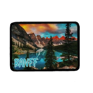 May include: A colorful embroidered patch with a scenic view of mountains and a lake. The text "BANFF" is written in blue on the bottom of the patch.