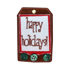 May include: A brown felt ornament with a red border and white fabric inside. The ornament has three green buttons and says "happy holidays!" in brown embroidery.