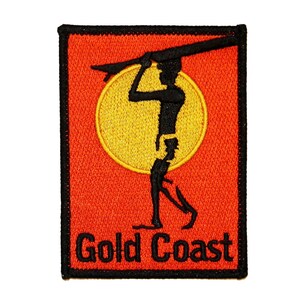 Può includere: Una toppa ricamata nera con la silhouette di una persona che tiene una tavola da surf davanti a un sole giallo su uno sfondo arancione. Il testo "Gold Coast" è scritto in nero sotto la silhouette.