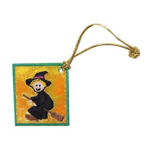 May include: A green and yellow square ornament with a witch riding a broomstick. The ornament has a gold cord attached to it.