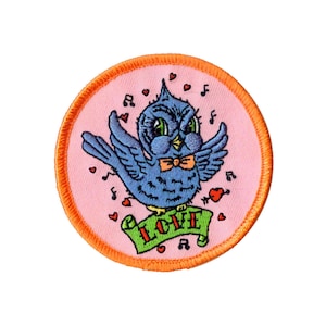 Peut inclure: Un patch brodé rose et orange avec un oiseau bleu chantant avec un nœud papillon et une bannière qui dit "LOVE".