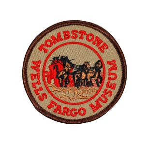 May include: A brown and tan circular patch with a red embroidered border. The text "Tombstone Wells Fargo Museum" is embroidered in red around the edge of the patch. A black and brown image of a stagecoach pulled by four horses is embroidered in the center of the patch.