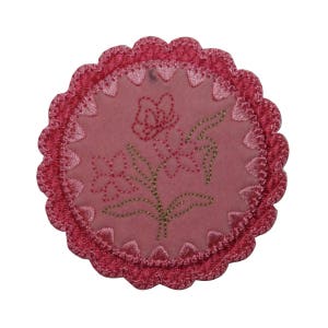 Può includere: Un sottobicchiere in tessuto rosa con bordo smerlato e un ricamo floreale in filo rosa e oro.
