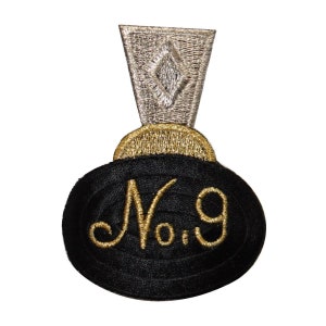 Può includere: Una toppa ricamata nera e dorata con il numero "No. 9" in un carattere dorato. La toppa ha un pezzo a forma di diamante argentato nella parte superiore.