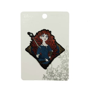 May include: A red-haired Disney princess, Merida, from the movie Brave, is depicted on an embroidered iron-on patch. She is holding a bow and arrow and is wearing a blue dress with a brown leather belt. The patch is on a white card with the Disney logo and the words "Brave" and "Iron-On Patch".