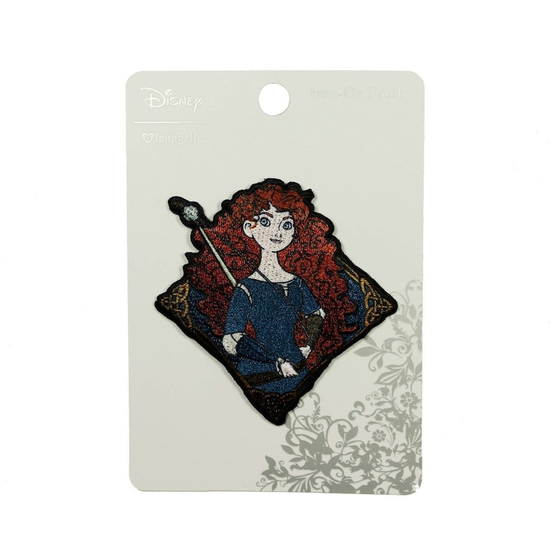 May include: A red-haired Disney princess, Merida, from the movie Brave, is depicted on an embroidered iron-on patch. She is holding a bow and arrow and is wearing a blue dress with a brown leather belt. The patch is on a white card with the Disney logo and the words "Brave" and "Iron-On Patch".