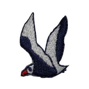 May include: A blue and white embroidered patch of a seagull in flight. The seagull has a red beak and white wings.