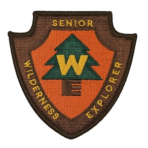 May include: A brown embroidered patch with a green pine tree and the letter "W" in yellow. The text "Senior Wilderness Explorer" is written around the tree in yellow.