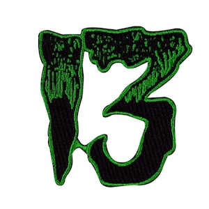 May include: Embroidered patch featuring the number 13 in a stylized, dripping font. The number is black with a vibrant green outline and interior details. The patch is designed for application on clothing or accessories.